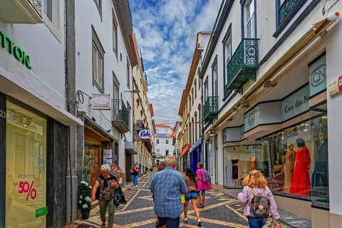 People walk through a busy shopping street with modern shops and historic buildings under a bright