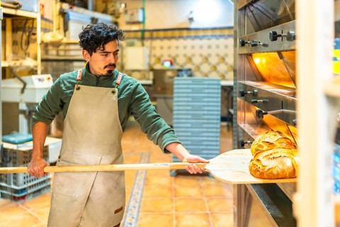 Latin young man using a wooden peel to remove baking bread from traditional oven
