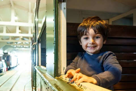 A young boy is smiling and looking out a window of a train. The train is in a station and the boy
