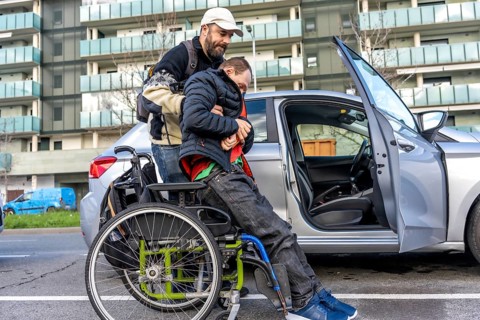 Man assisting disabled friend in getting out of a car parked on an urban street, showcasing the