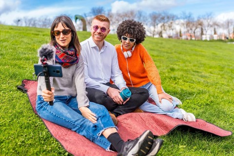 Three content creators sitting on a blanket in a park, recording a video blog using a smartphone