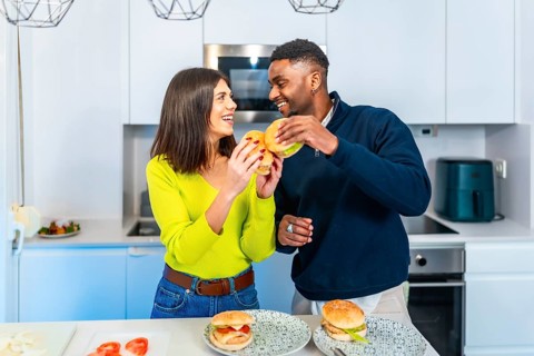 Cheerful couple enjoying a meal together, holding burgers and smiling in their contemporary kitchen