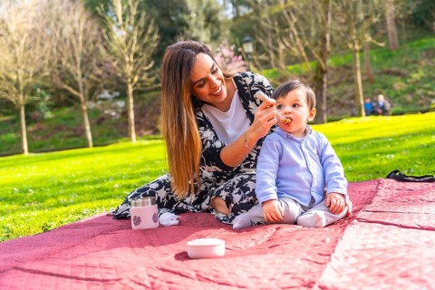Hispanic mother feeding her baby son solid food with a spoon while having picnic in a park,