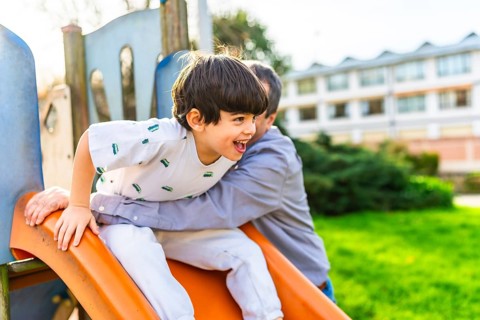 Grandfather holding grandson sliding down an orange slide at playground, having fun together on a