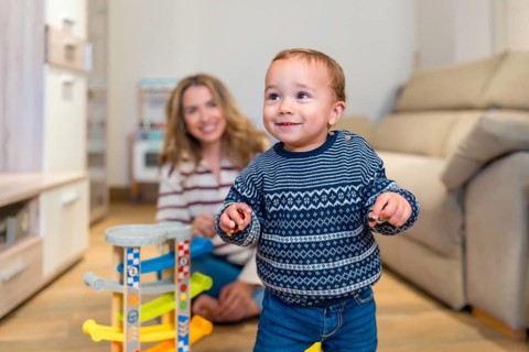 Happy baby boy learning to walk in living room with his mother watching and smiling, playing with a