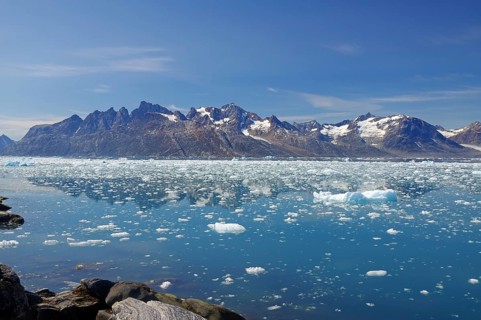 Icebergs floating in crystal clear water with mountains in the background under a blue sky, Knud