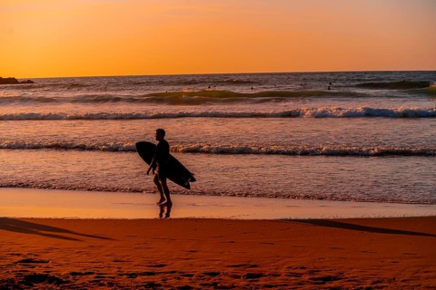 A man is walking on the beach with a surfboard. The sky is orange and the water is calm