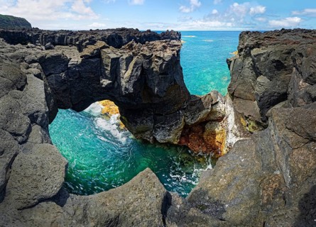 Panoramic view of rugged lava rock formations with a natural arch over turquoise sea under blue