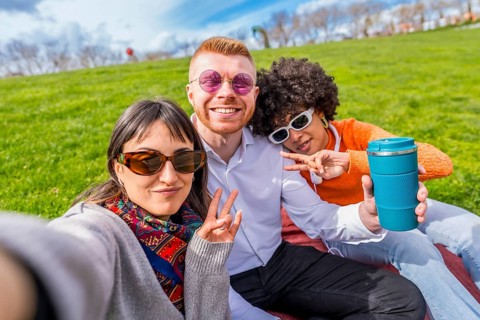 Three happy multi ethnic friends are taking a selfie in a park, gesturing victory signs and showing