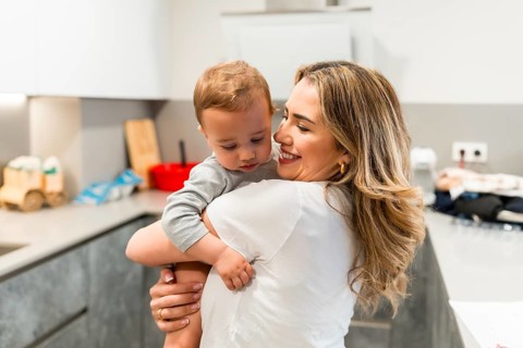 Smiling mother holding her baby son in a home kitchen, enjoying a tender moment of affection and