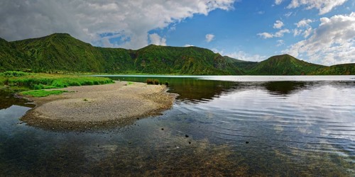Calm lake surrounded by green mountains under a partly cloudy sky, crater lake Lagoa do Fogo,