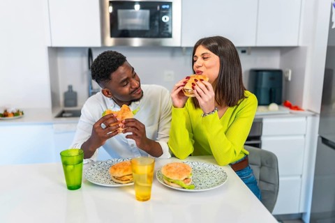 Young multiethnic couple eating burgers in their modern kitchen, enjoying a meal together
