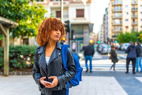 Latin young woman carrying bag in the city holding phone and looking at directions