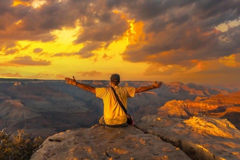 A young man sitting with his hands open and wearing a yellow shirt at Sunset at the Powell Point of