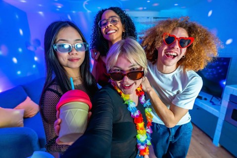 Four diverse friends enjoying a home disco party, taking a selfie with colorful lights, drinks, and