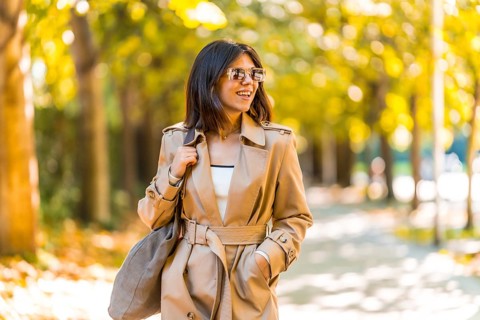 A woman wearing a tan trench coat and sunglasses walks down a sidewalk. She is smiling and she is