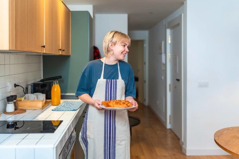 Smiling woman wearing apron carrying plate of fresh croissants in contemporary kitchen, enjoying