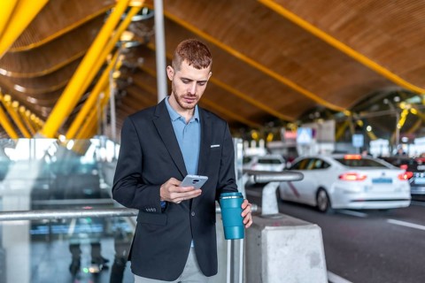 Young businessman using smartphone and holding reusable coffee cup while waiting for taxi or