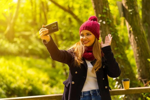 Young man in a black jacket, scarf and red woolen hat enjoying in an autumn park, waving on a video
