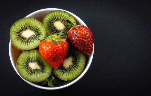 Sliced kiwis and strawberries in a white bowl with a black background