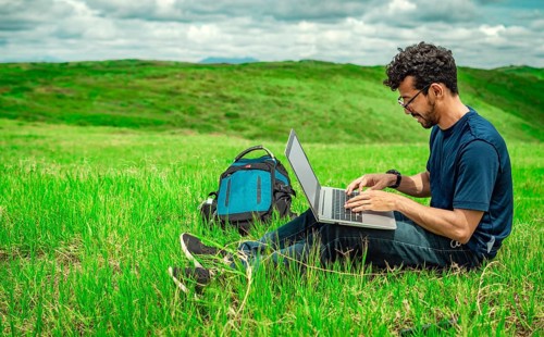 Close up of man sitting on green grass working with his laptop. Smiling freelancer guy working from