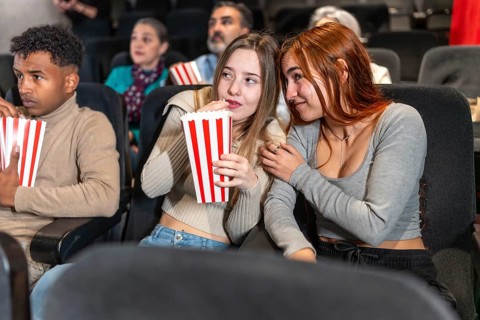 Two friends are whispering and eating popcorn while watching a movie at the cinema, surrounded by