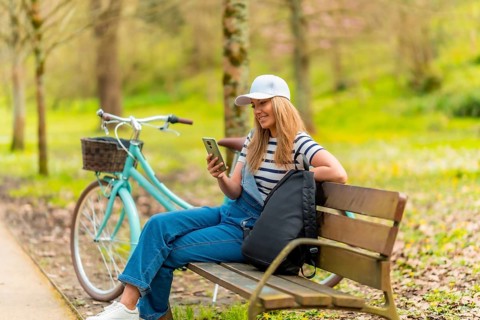 Young woman is using her smartphone while sitting on a bench in a park, with her bicycle parked