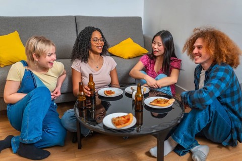 Four young friends sitting around a table, drinking beer and eating pizza, enjoying time together
