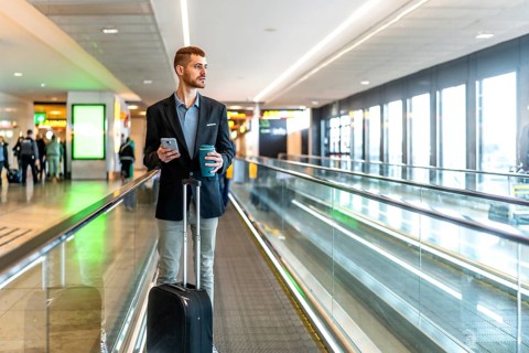 Businessman is walking on moving walkway at airport while using smartphone and holding coffee