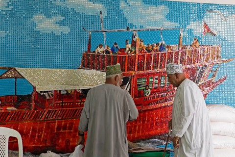 Omani men in dishdashas in front of the wall mosaic of a ship in Muttrah Fish Market, Muscat, Oman,