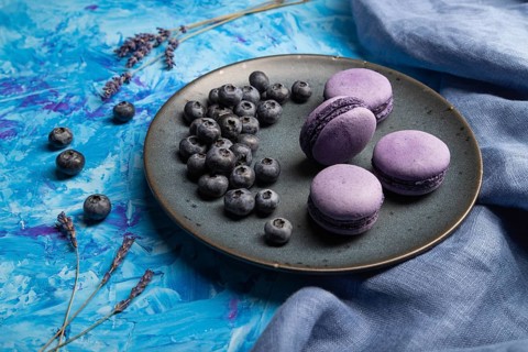 Purple macarons or macaroons cakes with blueberries on ceramic plate on a blue concrete background