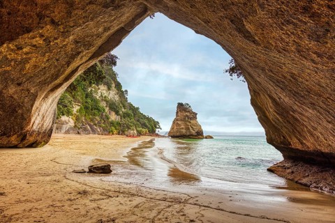 Looking out of a cave onto a rock formation and a quiet beach, Cathedral Grove, Coromandel, New