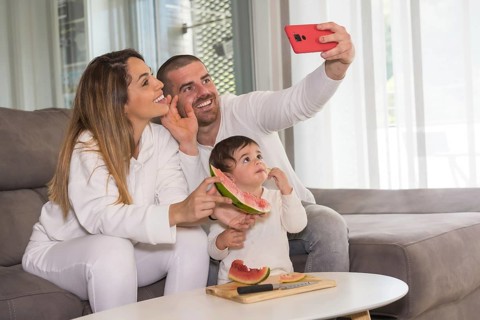 Family of three waving at camera taking a selfie while eating watermelon sitting on the sofa at