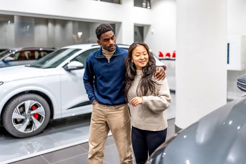 Happy multiethnic couple buying a new electric car at the dealership, embracing and choosing their