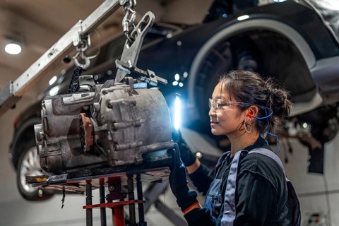 Young woman working as mechanic inspecting gearbox illuminated with flashlight in car workshop