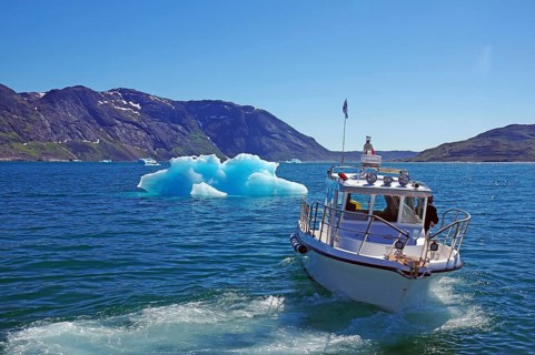 Boat sailing on a blue sea with iceberg and mountains in the background under a sunny sky, small