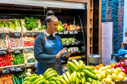 Young saleswoman using digital tablet and managing organic food in supermarket, she's wearing an