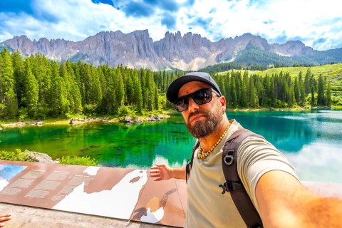 Tourist taking a selfie at lake carezza, with the stunning latemar massif and picturesque dolomites