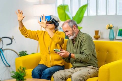 Senior woman using virtual reality googles next to her husband sitting on the sofa using mobile