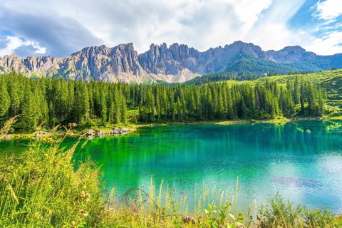 Emerald waters reflecting the latemar group and surrounding pine forest under a blue sky with