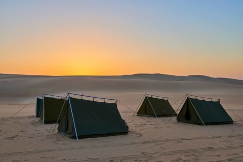 Four tents stand in a vast desert at dusk, surrounded by sand dunes under an orange-coloured sky,