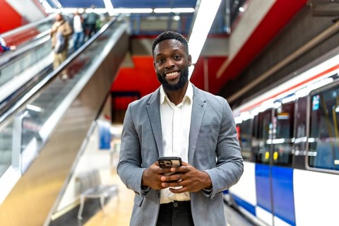 Young black businessman using smartphone while waiting for subway train in station, with escalator