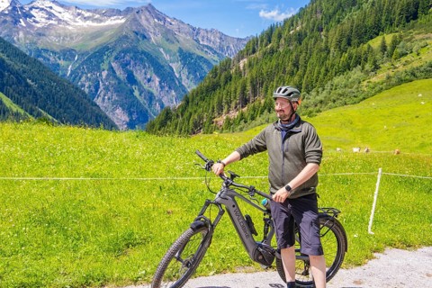 Man in sportswear standing next to his bicycle on a green meadow, surrounded by mountains and blue