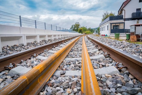A railway track runs through a landscape with buildings in the background, under a cloudy sky,