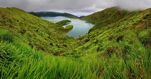 Natural landscape with a green slope and a lake surrounded by hills and a cloudy sky, crater lake