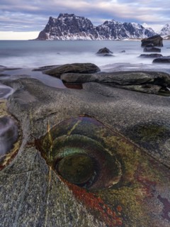 Rocks on the beach, rock formation Eye of Utakleiv, or Dragon's Eye, snow-capped mountains behind,