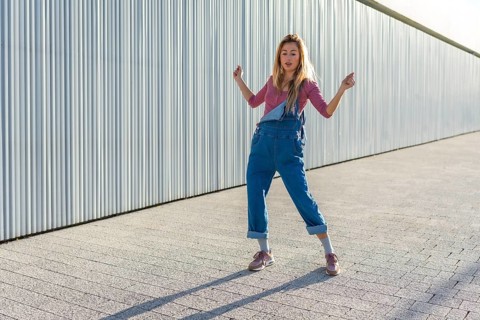 Blonde dancer performing urban dance moves on a paved area against a corrugated metal wall,