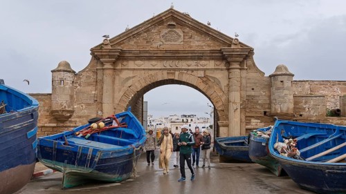 People walking through a stone arch, flanked by blue fishing boats, under a cloudy sky, Essaouira,