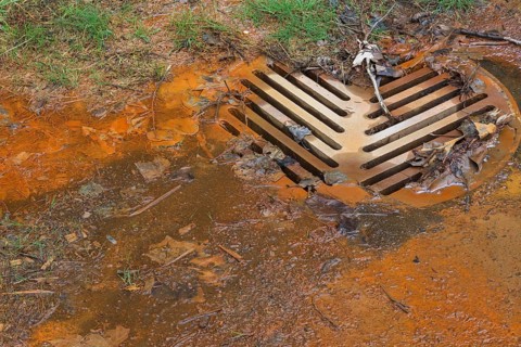 Contaminated rust colored water draining into sewer system at edge of a grass lawn in spring,