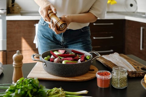 Close-up view of female hands sprinkle pepper from manual spice mill on vegetables in a baking pan